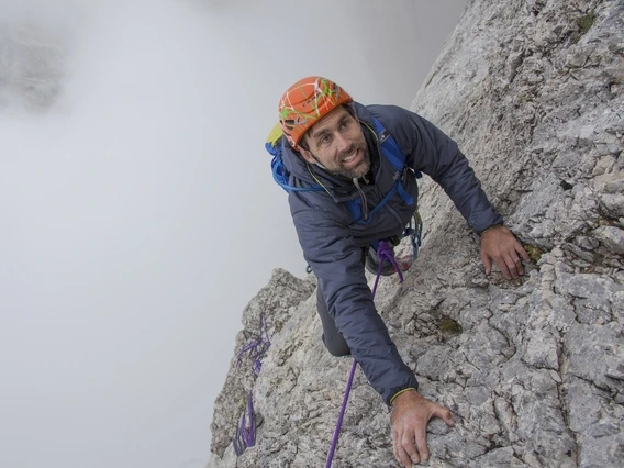 A climber in a gray jacket and bright orange helmet ascends a steep, rugged rock face using ropes and climbing gear. He looks up with determination, gripping the stone firmly as mist swirls around the mountain behind him.