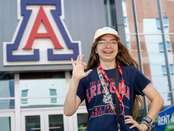 A young woman in a light colored hat and a blue shirt that says ARIZONA across it stands in front of a building with a large block A while holding up her right hand.