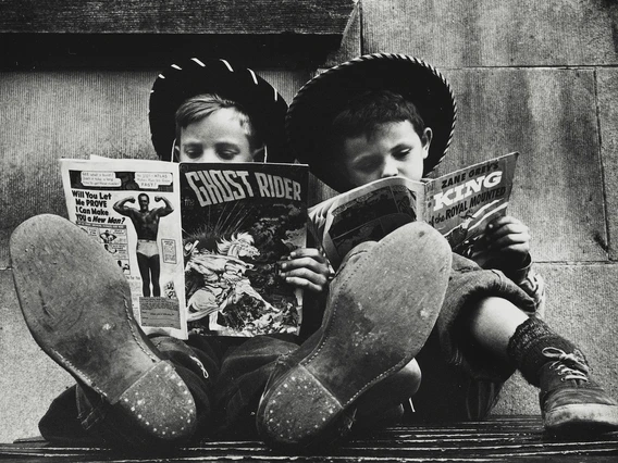 Two boys in cowboy hats sit side by side with their feet stretched out toward the camera, each reading a comic book. One holds The Ghost Rider while the other reads a Zane Grey western. The image is in black and white, highlighting their worn shoes and absorbed expressions.