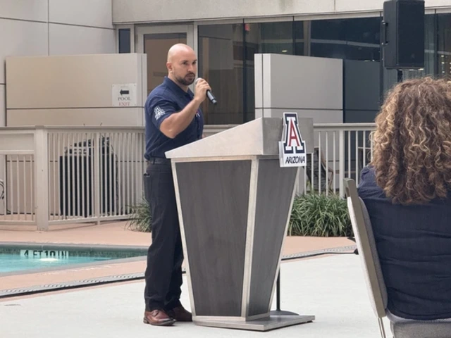 A man in a navy blue shirt speaking into a microphone at a podium. The podium is positioned near a swimming pool and features the University of Arizona logo with the word "ARIZONA" on it. In the background, there is a railing and plants adjacent to a modern building with large windows.