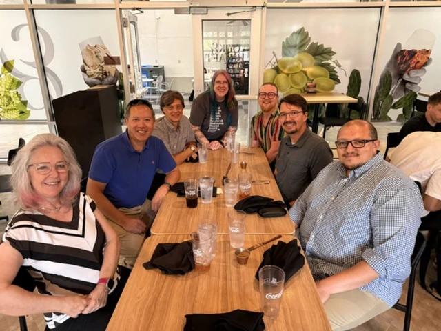 A group of seven people sit together at a long table in a restaurant, smiling at the camera during a casual lunch gathering. Drinks and utensils are on the table, and a bright interior with glass walls and food-themed images is visible in the background.