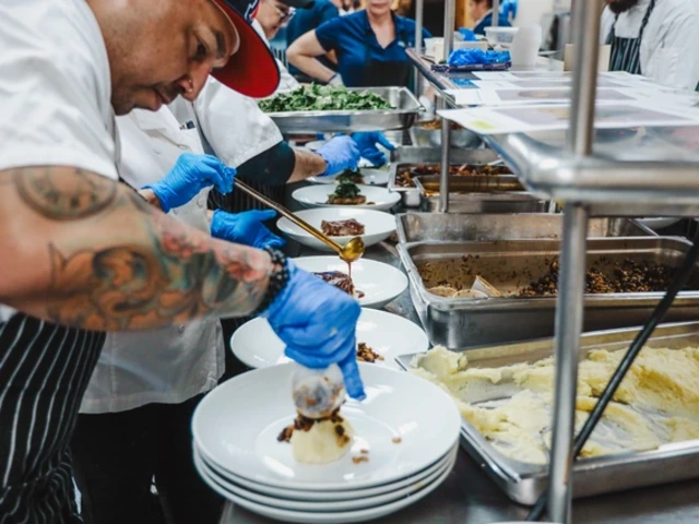 Chef in a red cap and tattooed arms plates dishes in a busy kitchen. Others assist with food preparation. Blue gloves worn, food on trays nearby.