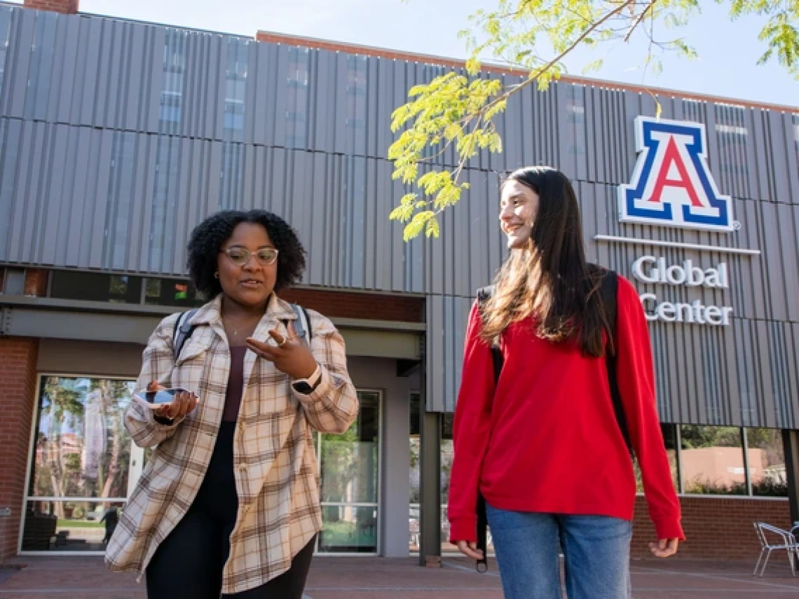 Two students walk and chat outside the University of Arizona Global Center. One student, wearing a plaid shirt and glasses, gestures while holding a phone, while the other, in a red sweatshirt and backpack, smiles. The modern building with the university's "A" logo is visible in the background.