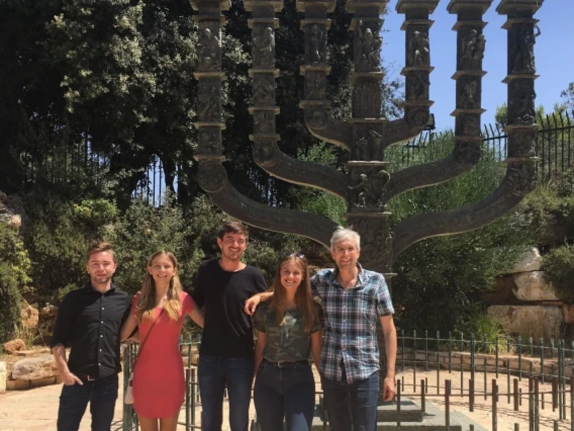 Five people standing together outdoors in front of a large bronze menorah sculpture. The group is smiling and dressed casually, with greenery and trees in the background. The sculpture features intricate designs and is surrounded by a low fence.