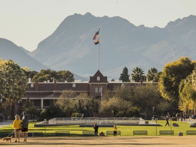 Wide view of the University of Arizona’s Old Main building in Tucson, Arizona, with the American and Arizona state flags flying above it, framed by tall palm trees and backed by mountains under a hazy sky. In the foreground, people walk dogs and take photos on the grassy mall, enjoying the sunny day.
