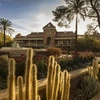 A historic brick building stands behind a garden filled with blooming red and purple flowers, tall cacti, and a fountain spraying arcs of water. Palm trees frame the scene as warm late-afternoon sunlight casts long shadows across the walkway.