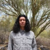 A man with long, wavy black hair stands outdoors in a desert-like environment with green trees and dry, rocky terrain in the background. He wears a buttoned-up, plaid long-sleeve shirt and has a serious expression on his face. The sky is bright and clear, with soft natural lighting casting shadows on the landscape