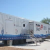 A large mobile veterinary clinic trailer against a clear blue sky. The trailer is white with blue accents and features images of animals, including a chicken and a dog. The left side displays the logo and text for the University of Arizona College of Veterinary Medicine. Metal stairs with railings lead to entrances on the side of the trailer. Various graphics, including cacti, are painted along the bottom.